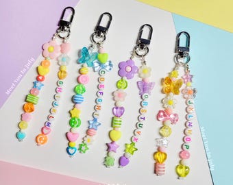 May include: Four colorful beaded keychains with silver clasps. Each keychain features a unique arrangement of beads, including flowers, butterflies, hearts, and letter beads. The keychains are displayed against a pastel background. The text "Meet You In July" is visible.
