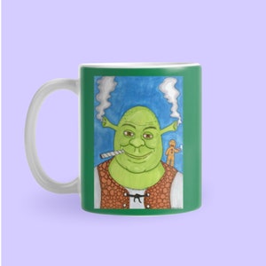 May include: A white ceramic mug with a green background featuring a cartoon illustration of Shrek smoking a joint with a gingerbread man. The mug has a green border and a white handle.
