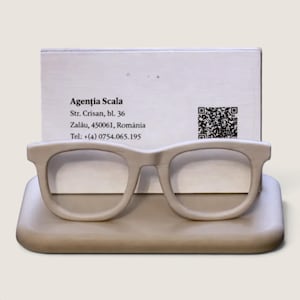 May include: A beige eyeglass holder with a rectangular base and a frame-shaped front. A white business card is inserted behind the frame, displaying the text Agenția Scala, Str. Crisan, bl. 36, Zalău, 450061, România, a phone number, and a QR code.