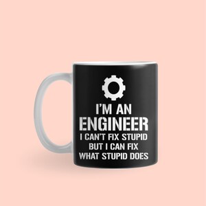 Puede incluir: Taza de café negra con asa blanca, con el texto blanco "I'M AN ENGINEER I CAN'T FIX STUPID BUT I CAN FIX WHAT STUPID DOES". Un engranaje blanco está encima del texto. La taza está sobre un fondo rosa claro.
