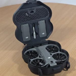 Op de afbeelding: Een grijze drone met zwarte propellers en een zwarte draagtas. De tas is open en toont de drone, twee batterijen en een klein gereedschap. De drone heeft een modern ontwerp.