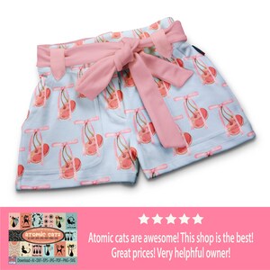 May include: A pair of light blue shorts with a pink bow tie and a pattern of pink cherries with pink bows. The shorts are made of a soft, comfortable fabric and have a relaxed fit.