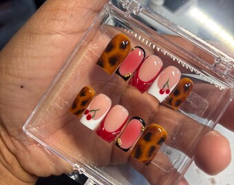 May include: A set of press-on nails in a clear plastic case. The nails feature various designs, including a leopard print, cherry accents, and French tips with red and black details. The nails are in a variety of shapes.