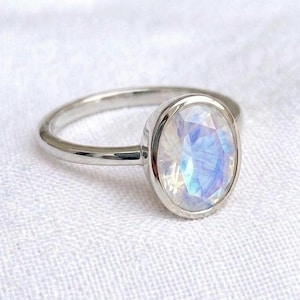 May include: A silver-toned ring featuring a single oval-shaped, faceted gemstone with a translucent, iridescent appearance that displays soft blue and white light reflections. The band is smooth, polished, and circular, holding the stone in a bezel setting.