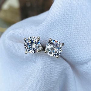 May include: A pair of dazzling, round-cut diamond stud earrings. The clear, faceted stones are set in a silver-coloured metal, highlighting their sparkle. The earrings are presented on a white fabric background.