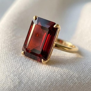 Puede incluir: Un anillo de oro con una gran piedra preciosa de color rojo intenso, de corte esmeralda rectangular, sujeta por cuatro garras. La banda es lisa y pulida, mostrando un diseño clásico que resalta el color vibrante y la claridad de la piedra.