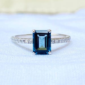 May include: A silver ring with a rectangular, dark blue gemstone as the centerpiece. The band is adorned with small, clear gemstones. The ring is set against a soft, white background, highlighting its elegant design.