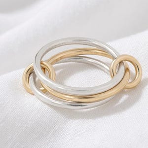 Puede incluir: Un anillo entrelazado de plata y oro. El anillo presenta tres bandas conectadas, con dos bandas plateadas y una dorada. El anillo está sobre un fondo blanco.