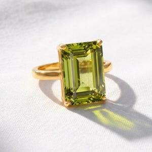 May include: A gold ring featuring a large, rectangular, emerald-cut lime green gemstone set in a four-prong mounting. The band is smooth and polished, showcasing a vibrant, translucent green stone that reflects light onto the surface below.