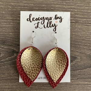 May include: A pair of dangle earrings with a gold and burgundy leather design. The earrings are displayed on a white card with the text "designs by Lilly" and a small pink heart.