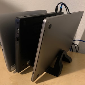 May include: Three laptops stacked on top of each other on a wooden surface. The laptop on the bottom is silver and has a black stand. The middle laptop is black and the top laptop is silver.