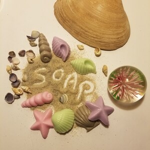 May include: A collection of handmade soap bars shaped like seashells and starfish, arranged on a bed of sand with the word "SOAP" written in the sand. The soaps are in shades of pink, purple, green, and brown.