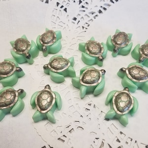 May include: A collection of twelve green and silver turtle shaped wax melts. The wax melts are arranged on a white doily.