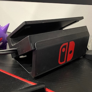 May include: A black Nintendo Switch dock with a red Nintendo logo. A purple Gengar Pokemon figurine is in the foreground.