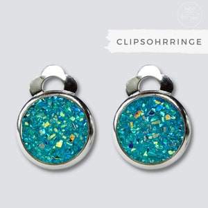 Könnte beinhalten: Ein Paar silberne Ohrclips mit einem runden, türkisblauen Glitzerdesign. Die Ohrringe haben oben ein blumenförmiges Design.