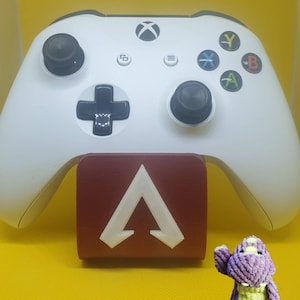 May include: A white Xbox controller with a red stand featuring a white Apex Legends logo. A purple plush dinosaur toy is in the foreground.