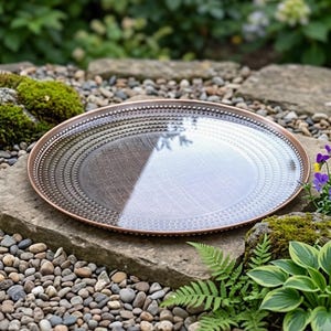 May include: A round, shallow tray with a metallic copper-toned rim and a textured surface featuring a gradient pattern that transitions from a dark, speckled gray on one side to a lighter, reflective silver finish on the opposite side.