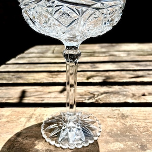 May include: A clear, vintage champagne coupe glass with a detailed, cut-glass design. The bowl has a diamond and starburst pattern, supported by a slender stem and a star-shaped base. The glass sits on a wooden surface.