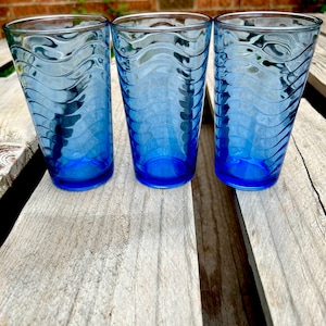 May include: Three blue glass tumblers with a wavy design. The glasses are sitting on a wooden surface.