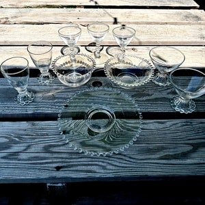 May include: A set of clear glass dessert dishes with beaded edges. The dishes are arranged on a wooden surface with a larger beaded glass serving plate in the center.
