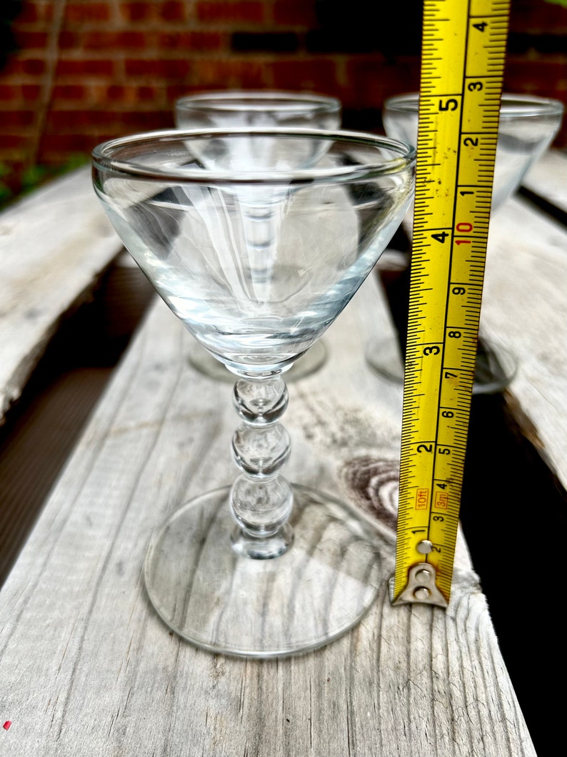 K&ouml;nnte beinhalten: Drei klare Schnapsgl&auml;ser aus Glas mit einem perlenbesetzten Stiel. Die Gl&auml;ser stehen auf einer Holzoberfl&auml;che mit einem gelben Ma&szlig;band.