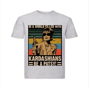 Op de afbeelding: Lichtgrijs t-shirt met een retro graphic van een vrouw met een zonnebril die een fles vasthoudt. De graphic bevat de tekst "IN A WORLD FILLED WITH KARDASHIANS BE A PATSY!" in vintage stijl.