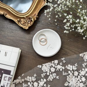 Peut inclure: Un petit plat rond blanc avec le mot "for" gravé. Deux bagues, une argentée et une en or rose, sont à l'intérieur. Un miroir doré orné et des fleurs blanches sont en arrière-plan.