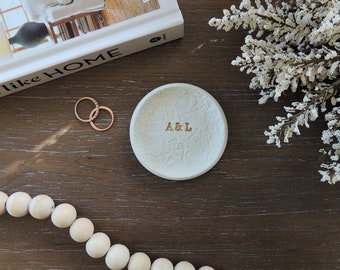 May include: A white ceramic ring dish with a floral pattern and the initials "A & L" in gold lettering. The dish is on a dark brown wooden surface with a string of wooden beads and a book.