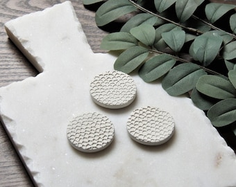 May include: Three small, round, white ceramic dishes with a textured, scale-like pattern. The dishes are arranged on a white marble surface.