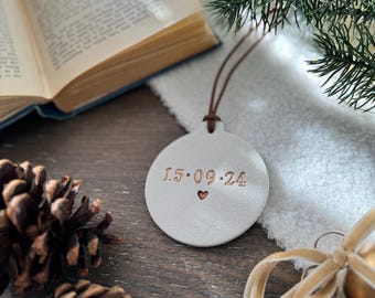 May include: A white, round Christmas ornament with the date "15.09.24" and a heart symbol. The ornament hangs by a brown leather cord. The background includes an open book, pine cones, and a sprig of evergreen.