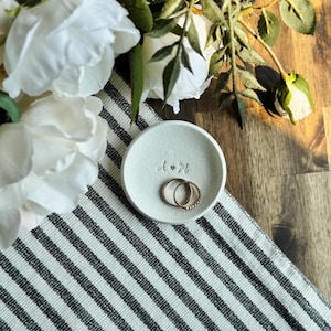 May include: A small, round, off-white ring dish with the initials "A & M" engraved on it. Two gold rings rest inside the dish. The dish sits on a black and white striped cloth, with white flowers and greenery in the background.