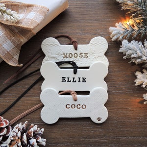 May include: Three white ceramic dog bone ornaments with brown leather cords. The bones are stacked on top of each other. The top bone has the name "MOOSE" written on it. The middle bone has the name "ELLIE" written on it. The bottom bone has the name "COCO" written on it.