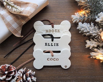 May include: Three white ceramic dog bone ornaments with brown leather cords. The bones are stacked on top of each other. The top bone has the name "MOOSE" written on it. The middle bone has the name "ELLIE" written on it. The bottom bone has the name "COCO" written on it.
