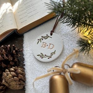 May include: White ceramic ornament with a gold 'B & D' monogram surrounded by green leaves and red berries. The ornament is hanging from a brown cord.