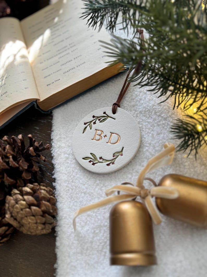 May include: White ceramic ornament with a gold 'B & D' monogram surrounded by green leaves and red berries. The ornament is hanging from a brown cord.