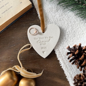 May include: A heart-shaped ornament with the text "The Year We Got Engaged" and the year "2025". A rose gold engagement ring is attached. The ornament hangs from a brown ribbon. Gold bells and pine cones are also visible.