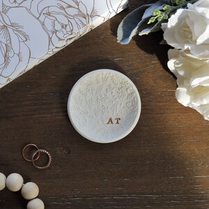 May include: White ceramic ring dish with a floral design and the initials "AT" in gold lettering.