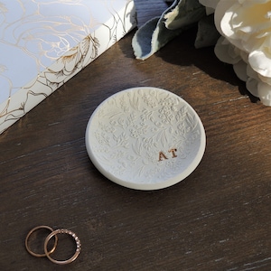 May include: White ceramic ring dish with a floral design and the initials "AT" in gold lettering. Two gold rings are on a wooden surface.