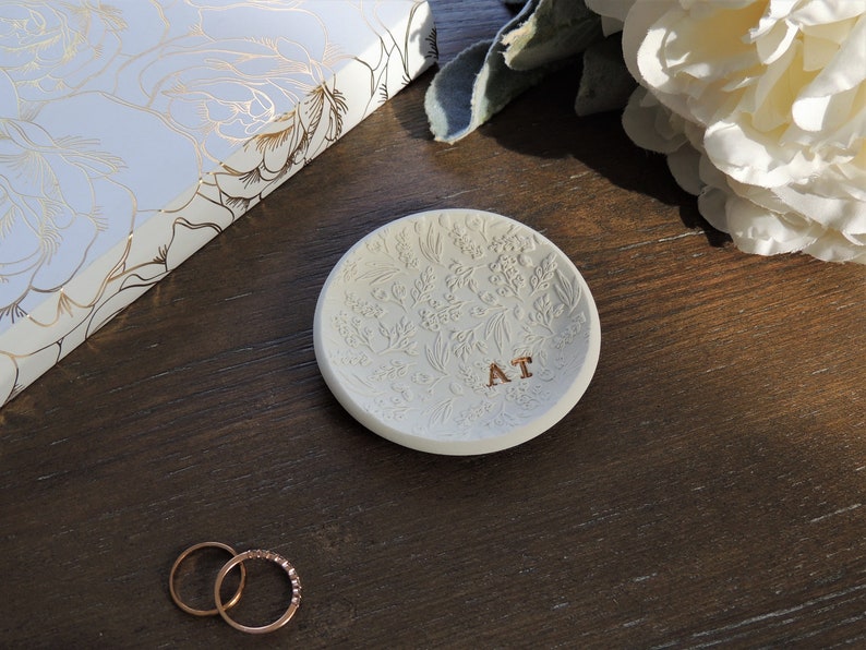 May include: White ceramic ring dish with a floral design and the initials "AT" in gold lettering. Two gold rings are on a wooden surface.