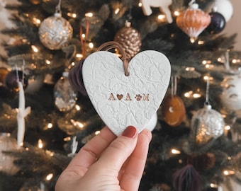 Pode incluir: Adorno de coração branco de cerâmica com um design floral e o texto "A ♥ A & M" em letras douradas.