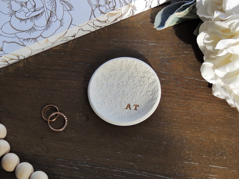 May include: White ceramic ring dish with a floral design and the initials "AT" in gold lettering. The dish is on a dark brown wooden surface.
