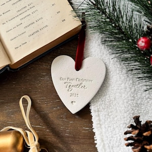 May include: A heart-shaped Christmas ornament with the text "Our First Christmas Together 2025" is displayed. The ornament hangs from a red ribbon. The background includes a book, a white towel, and pine branches.