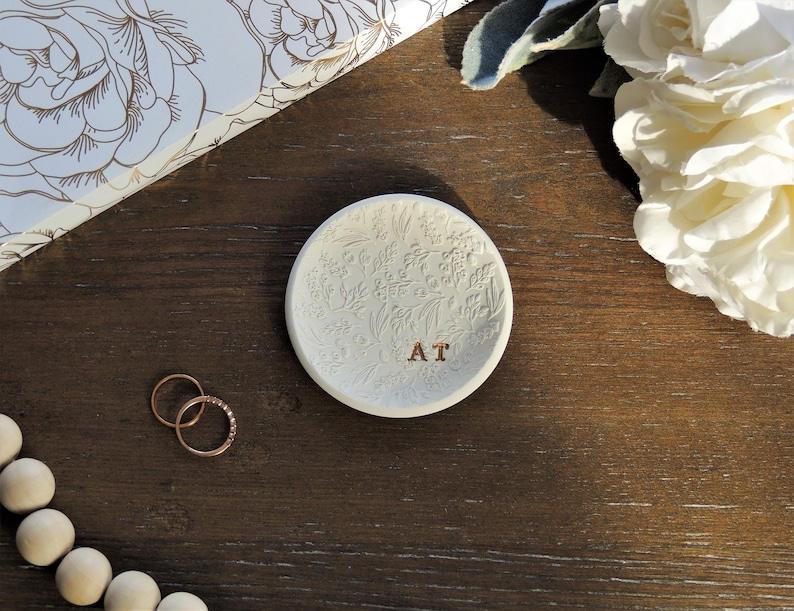 May include: White ceramic ring dish with a floral design and the initials "AT" engraved in the center. The dish is on a wooden surface.