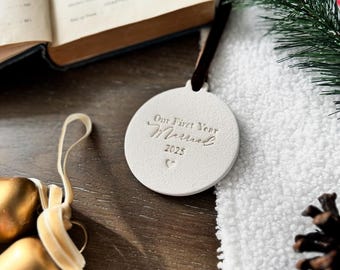 May include: A white, round Christmas ornament with the inscription "Our First Year Married 2025" in elegant script. The ornament hangs from a dark brown ribbon. Gold-colored bells and a pine branch are also visible.