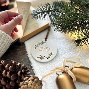 May include: White ceramic ornament with a green floral wreath and the initials "B & D" in gold lettering. The ornament is hanging from a brown cord and is surrounded by pine branches, pine cones, and gold bells.