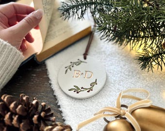 May include: White ceramic ornament with a green floral wreath and the initials "B & D" in gold lettering. The ornament is hanging from a brown cord and is surrounded by pine branches, pine cones, and gold bells.