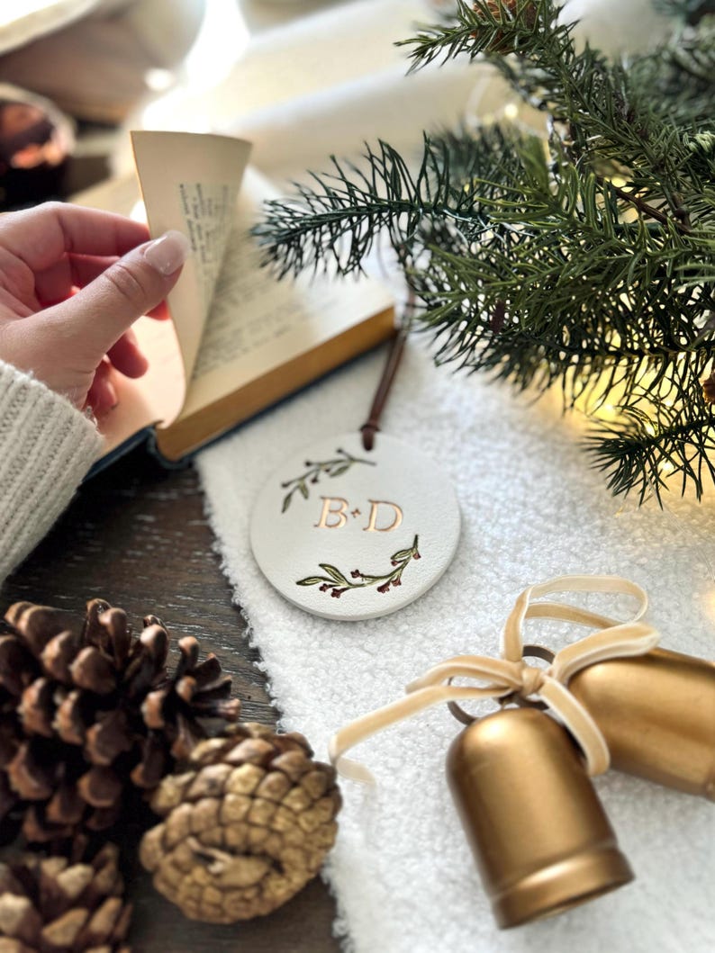 May include: White ceramic ornament with a green floral wreath and the initials "B & D" in gold lettering. The ornament is hanging from a brown cord and is surrounded by pine branches, pine cones, and gold bells.