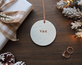 May include: White round ornament with a textured surface and the initials "V & M" engraved in gold. The ornament is hanging from a brown cord.