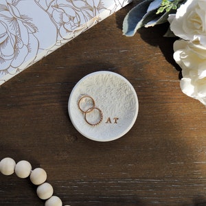 May include: White ceramic ring dish with a floral design and the letters "AT" inscribed on the bottom. Two gold rings are resting on the dish.