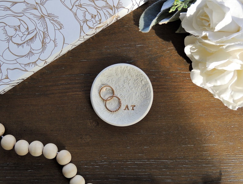May include: White ceramic ring dish with a floral design and the letters "AT" inscribed on the bottom. Two gold rings are resting on the dish.